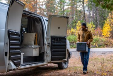 Camper carrying a portable power station beside a van among autumn trees
