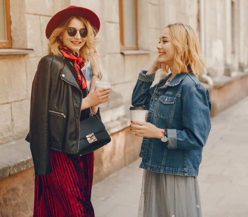 a young and stylish girl with light hair dressed in a white T-shirt and gray skirt, standing in the summer city and drinking a coffee with her girlfriend dressed in a black T-shirt and a red skirt
